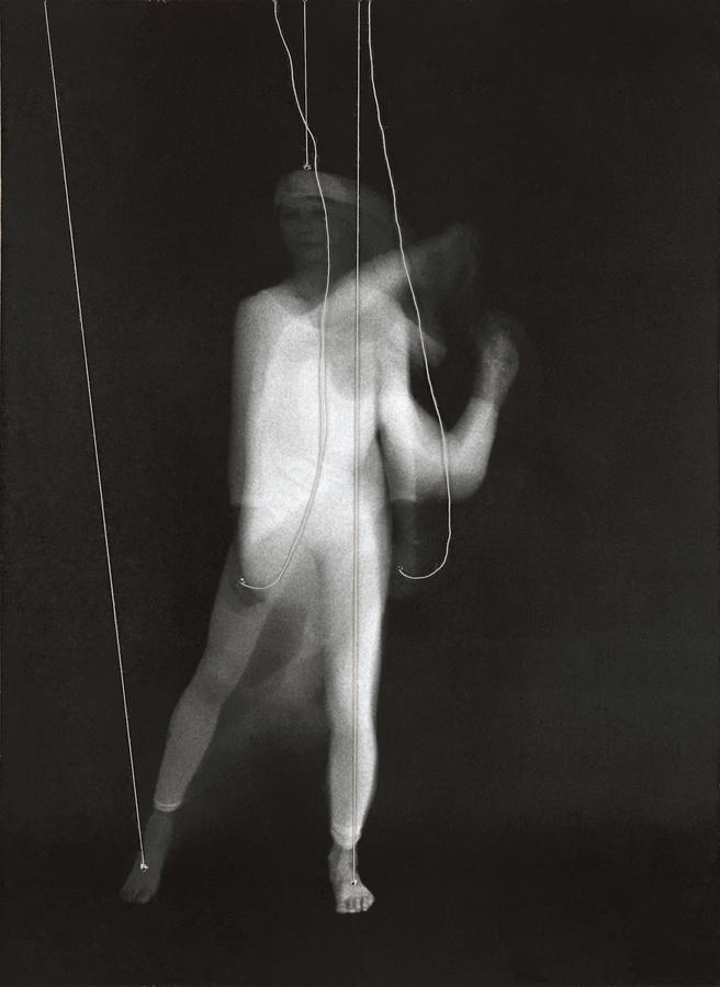 A blurry image of a woman in a white body suit with strings attached to her feet, hands, and head