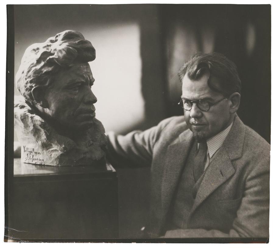 A man in a suit and circle glasses leaning his arm on a sculpture of a man’s head. Black and white photograph by Violet Keene Perinchief.