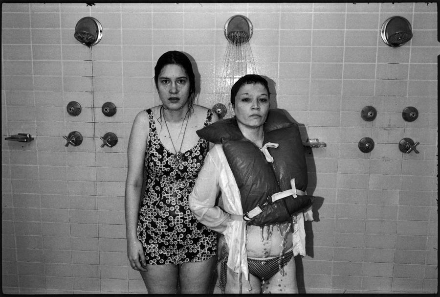 A black and white photograph of two women with serious expressions standing under a running shower, one in a full-piece bathing suit, and the other in a bikini, a white shirt and a life jacket.