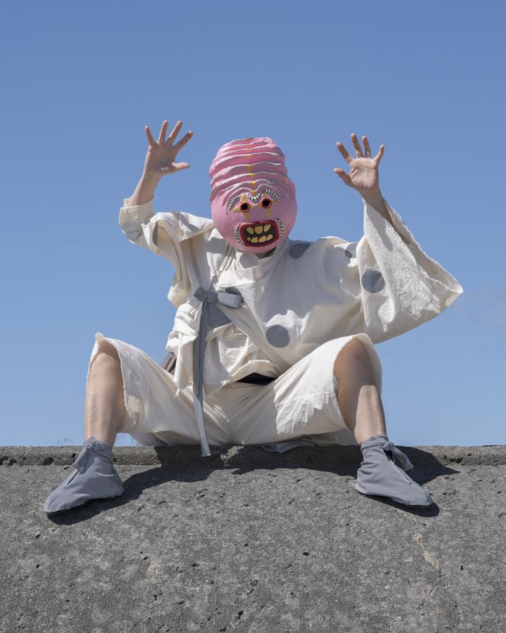 A figure in a white kimono-style outfit and wearing a pink mask holds up their arms in a menacing gesture while sitting casually on a concrete form.