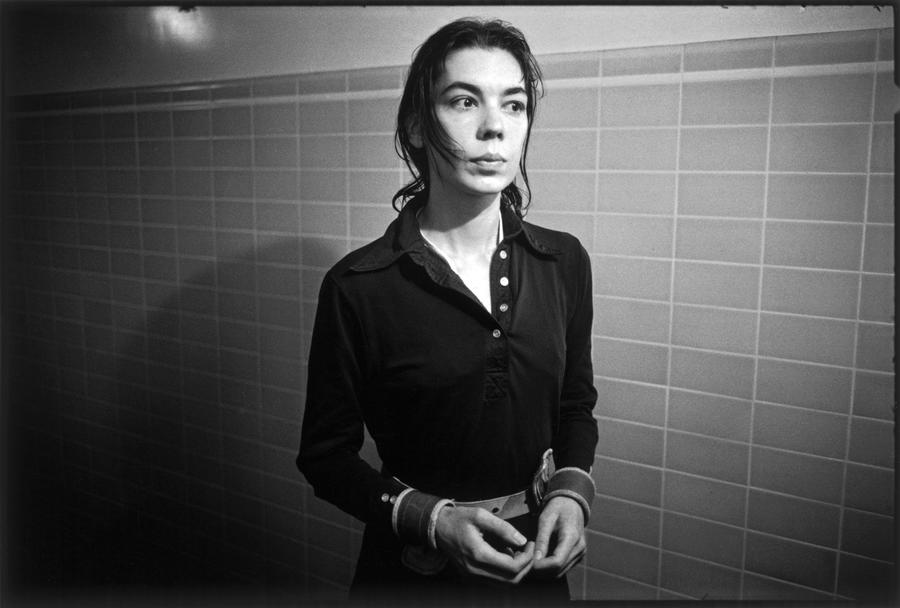 A black and white image of a woman in a black collared dress stands in a tiled hallway wearing hand restraints