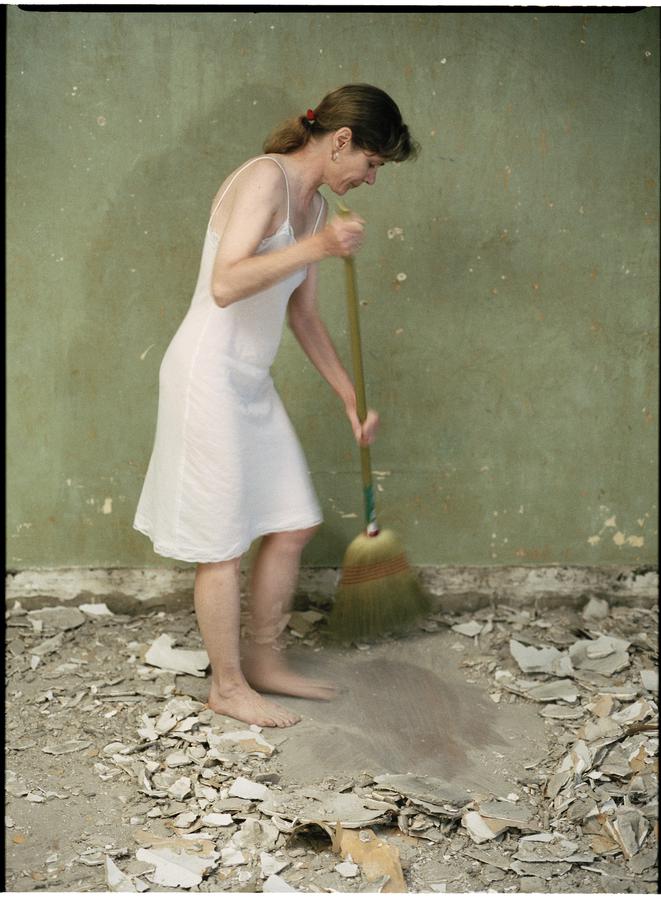 A woman in a white sleeveless dress and no shoes sweeps plaster off the floor in front of a green wall