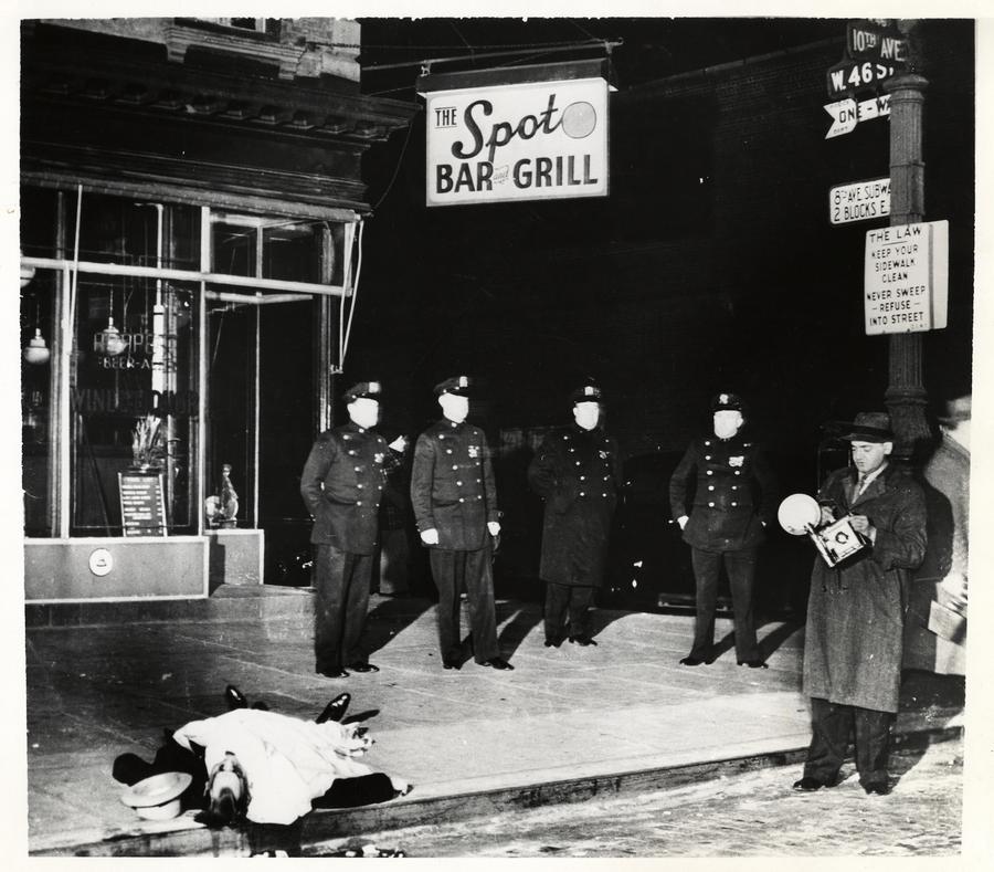 A group of police officers and a journalist gather around a dead body in front of The Spot Bar and Grill