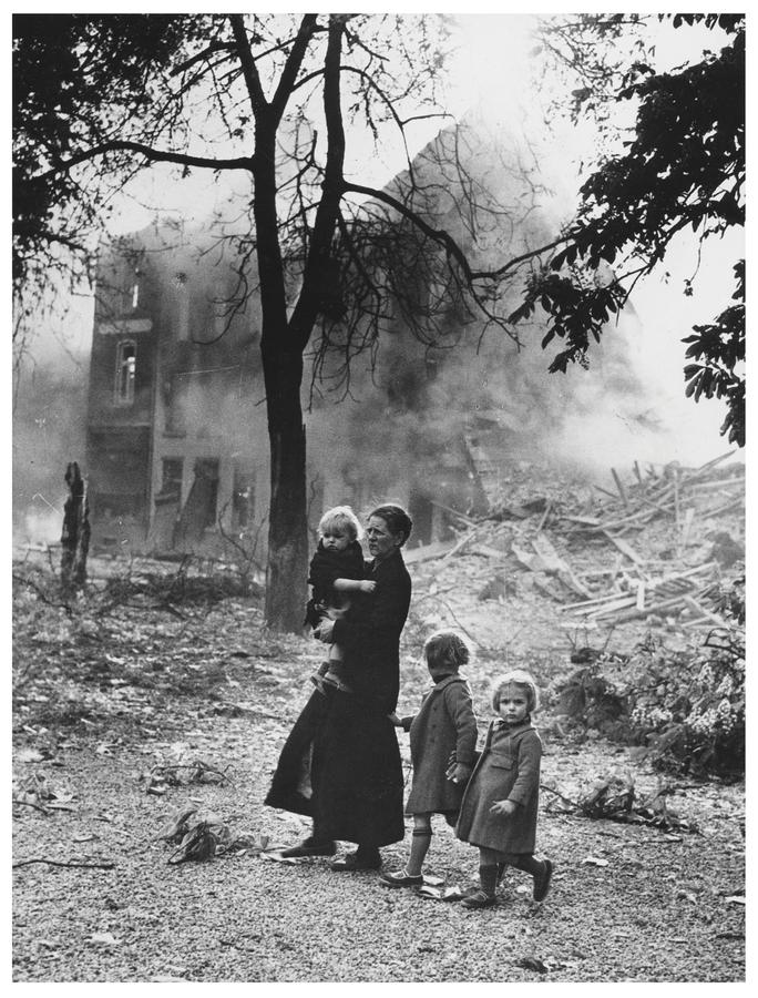 A scene of war, in which a mother and three children stand in front of a destroyed building