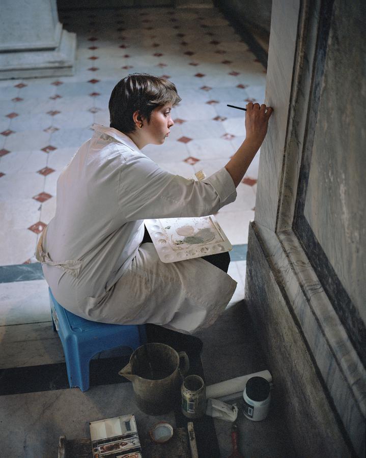 A woman seated on a stool next to a wall that she is painting with a small brush.