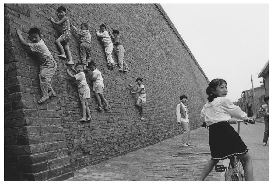 Kids climb a wall while a little girl rides a bike in the foreground.
