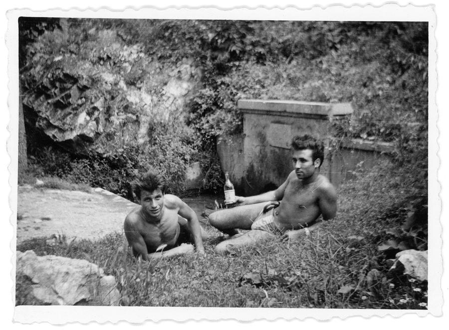 An old photograph of two shirtless men lying in a patch of grass, one of them holding a bottle