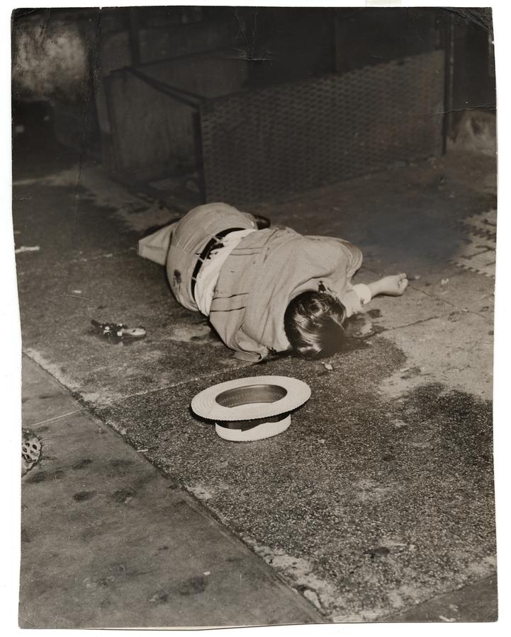 A dead man's body lying on the sidewalk, his hat upside down on the ground beside him