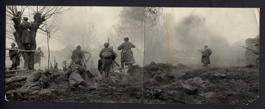 Two armed soldiers lay on sloped ground aiming their rifles, several more soldiers standing in front of them while another observes from the crevice of tree branches