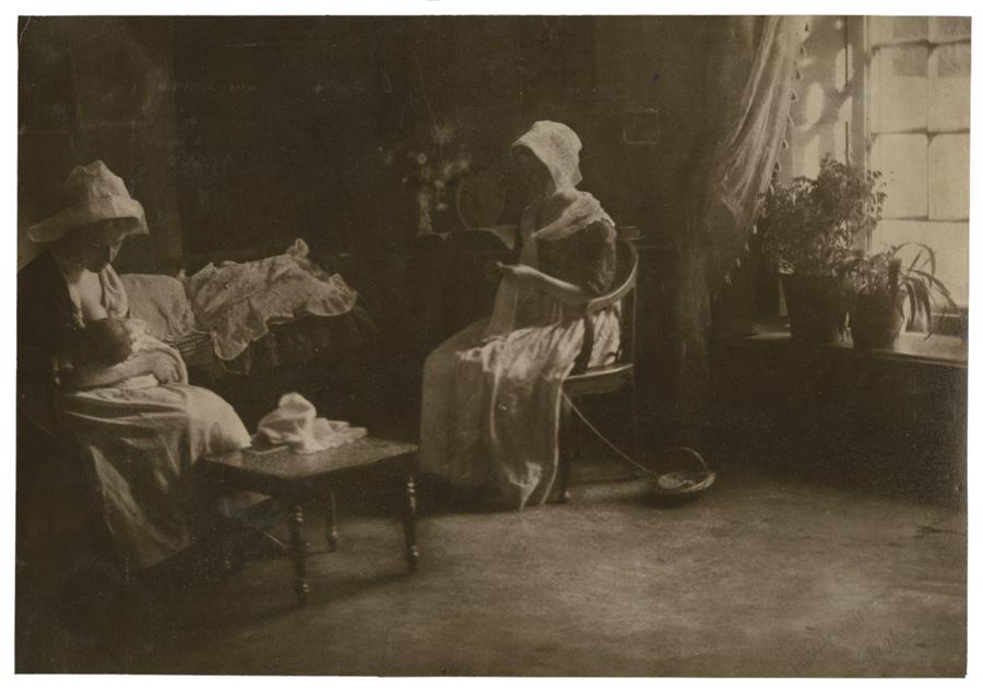 Two women sitting in a dimly lit living room, one knitting while the other holds a baby. Black and white photograph by Minna Keene.