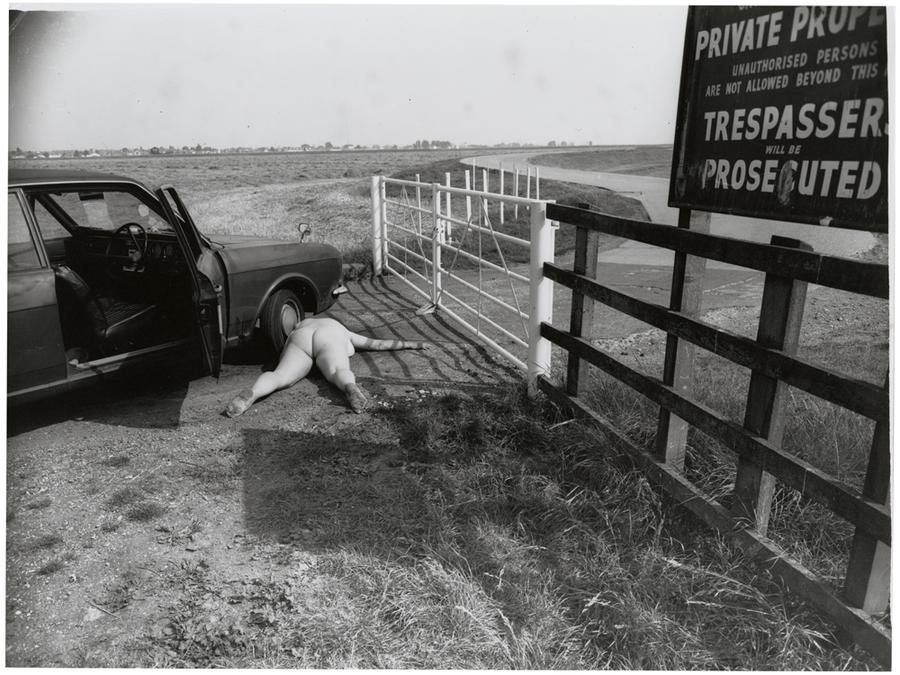 Nude person laying on the ground next to a car with open doors in front of a gated road.