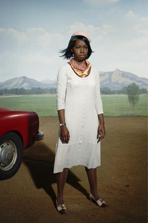 A woman in a white dress, pink hat, and thin scarf standing in front of a red car