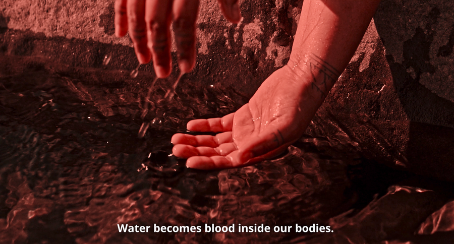 Red filter over still of two hands draped over stone. Caption reads "Water becomes blood inside our bodies."