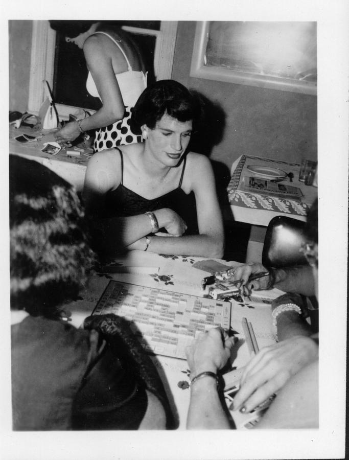 A group of men wearing dresses and jewelry play Scrabble