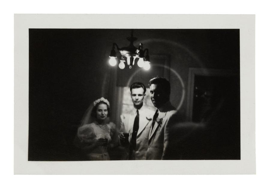 newlywed man and woman stand beside groomsman beneath bright overhead lightbulbs in a vignette of darkness