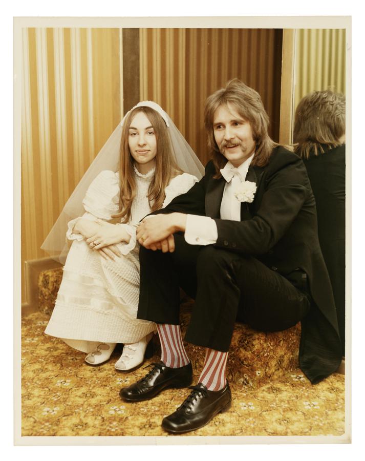 newlywed bride and groom sit on a patterned carpet floor. Bride wears a veil, groom has a mustache, a mullet and red striped socks.