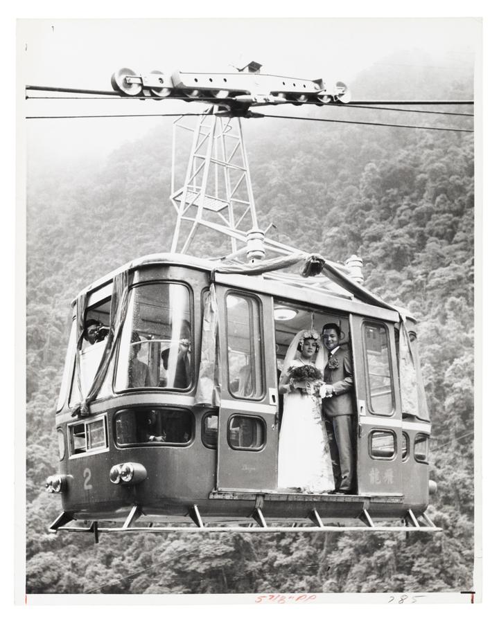 newly wed husband and wife stand in the open door of a cable car, shot from below in front of a mountain