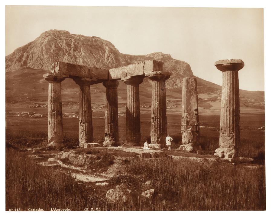 Two people in front of an acropolis in Corinth, Greece. The acropolis is located in a grassy field, with a mountain in the background.