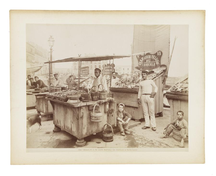 An oyster merchant pictured at a booth showcasing his wares. A number of other men and boys are pictured around the booth looking into the camera