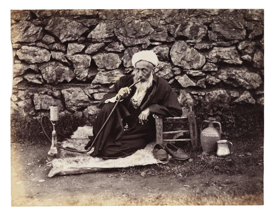 A seated man with a bear smokes a hookah by a rock wall.
