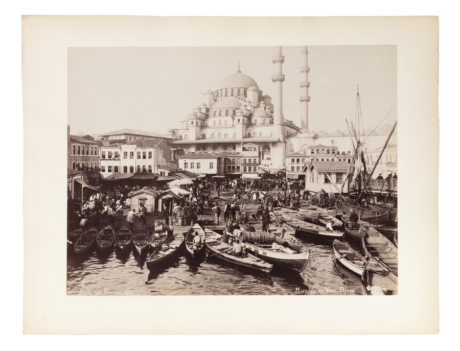 The photograph captures a bustling waterfront scene in Istanbul, centered around the Yeni Cami (New Mosque), a prominent Ottoman imperial mosque located in the Eminönü district near the Golden Horn.

In the foreground, the quay is crowded with small wooden rowboats and fishing vessels, some filled with goods, possibly fish or textiles. People are actively loading, unloading, or navigating the crowded shoreline, indicating a lively commercial hub or market area.

Behind the waterfront, a densely packed collection of Ottoman-era buildings leads the eye upward toward the monumental Yeni Mosque, with its grand central dome, semi-domes, and twin minarets rising into the sky. The mosque dominates the skyline and serves as both a religious and architectural focal point.

The photograph is sepia-toned and mounted on a white border, typical of late 19th-century albumen prints, and bears the studio mark: “Abdullah Frères, 663” in the bottom left, and
“Mosquée de Yeni-Djami” in the bottom right.