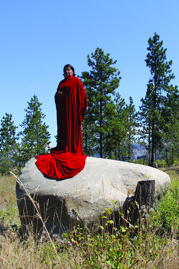 A woman in a long red cloak stands on top of a large rock