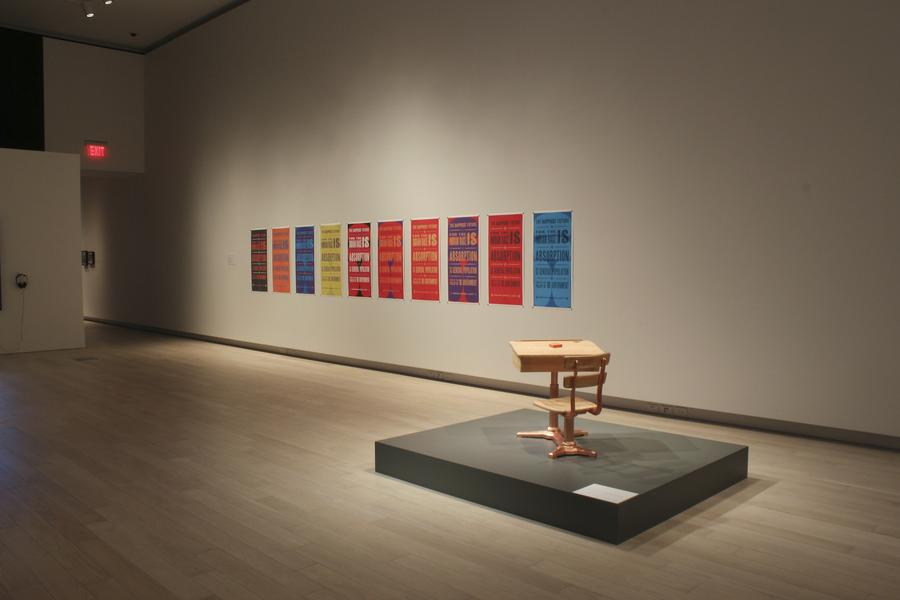 A series of colourful posters. An old school desk on a black platform in the center of the gallery.