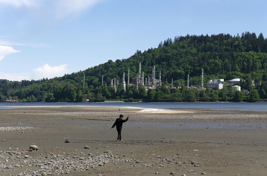 In the foreground, a boy dances on a beach by the water. An oil refinery is shown in the background in a forested area.