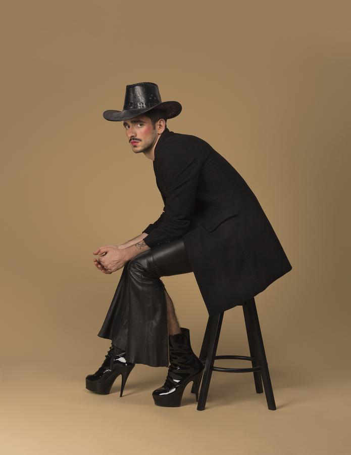 A young man posing on a stool wearing a black hat and high heels