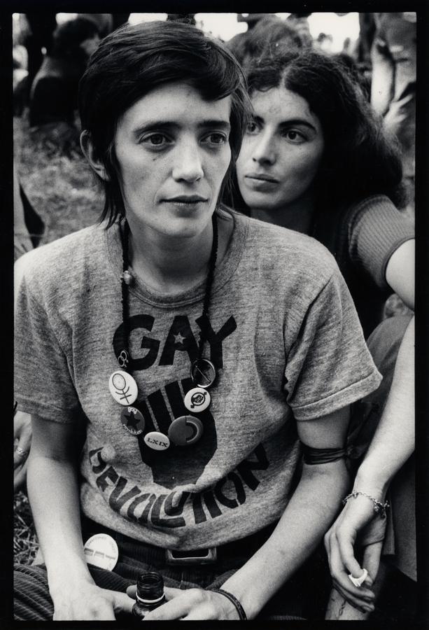 A woman with short hair wearing a shirt that says "gay revolution". Another woman sits behind her