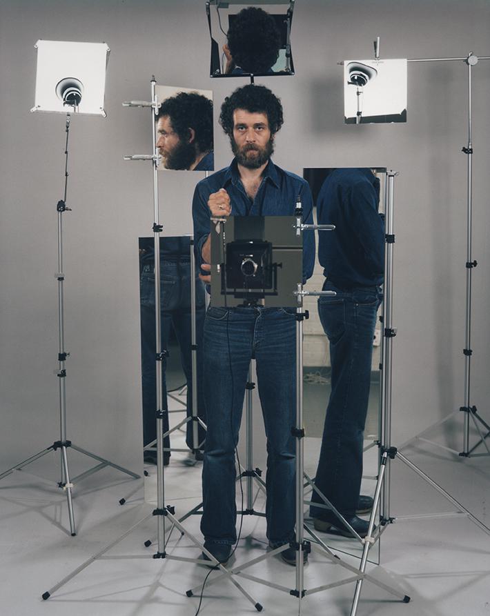A man with curly hair and a beard stands in the middle of a cluster of mirrors in a photo studio