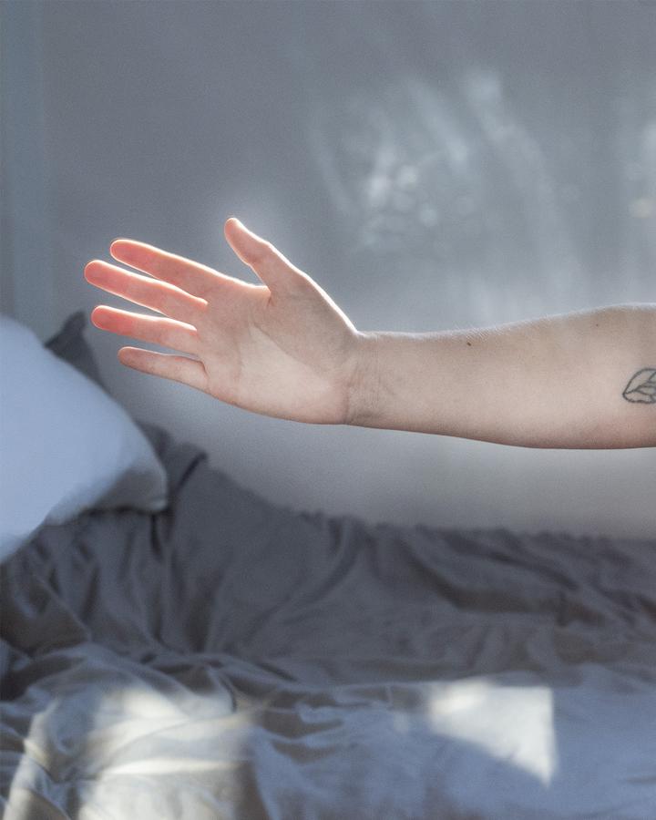 A photo of an outstretched hand and arm, with a small tattoo visible at the elbow. The arm is backlit and shown in front of a bed.