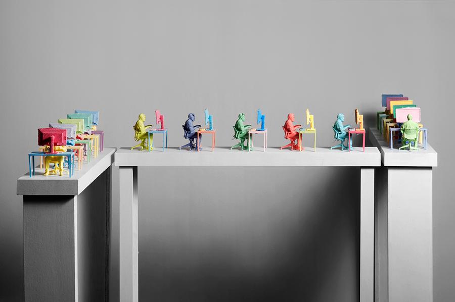 Multiple miniature colourful figurines of men sitting at a desk with a computer are posed on a beam
