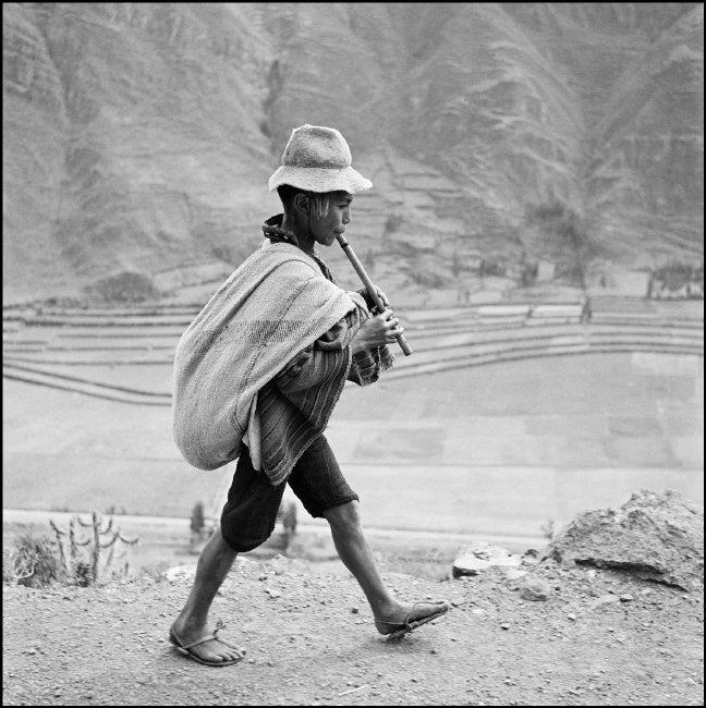 Young Peruvian boy wearing straw hat and rope sandals carrying a blanket sack on his back, walks in the foreground of a mountain scape while playing a flute.