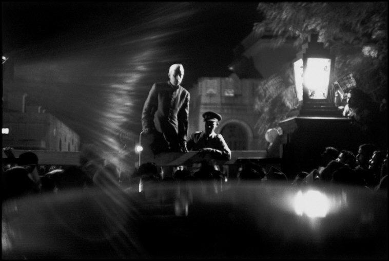 Two men standing on platform in front of a blurry crowd, illuminated only by a lamp post on a dark night.
