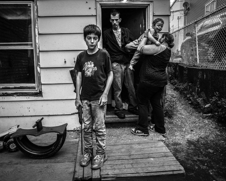 Black and white image of a boy holding a rifle stands in the foreground with his family behind him in front of a run down home. A man, woman and child make up his family.