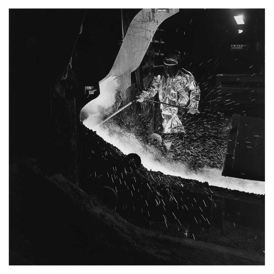 A black and white photo of a miner wearing protective gear working with a furnace.