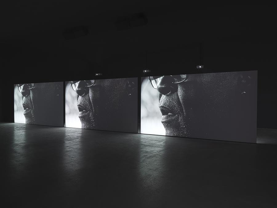 3 large screens in an empty room show the same image repeated, a close up of an open mouth