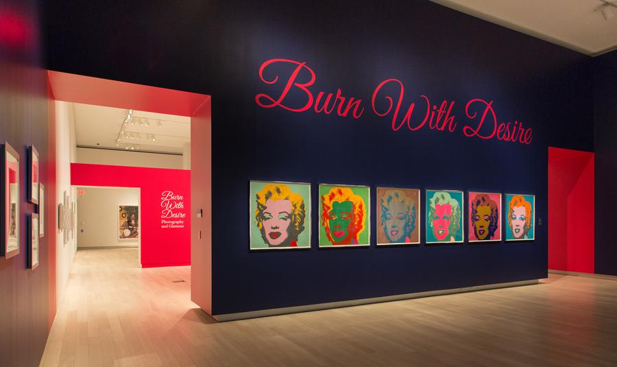 6 brightly coloured portraits of Marilyn Monroe on a deep blue wall, pink script above reads "Burn with desire"