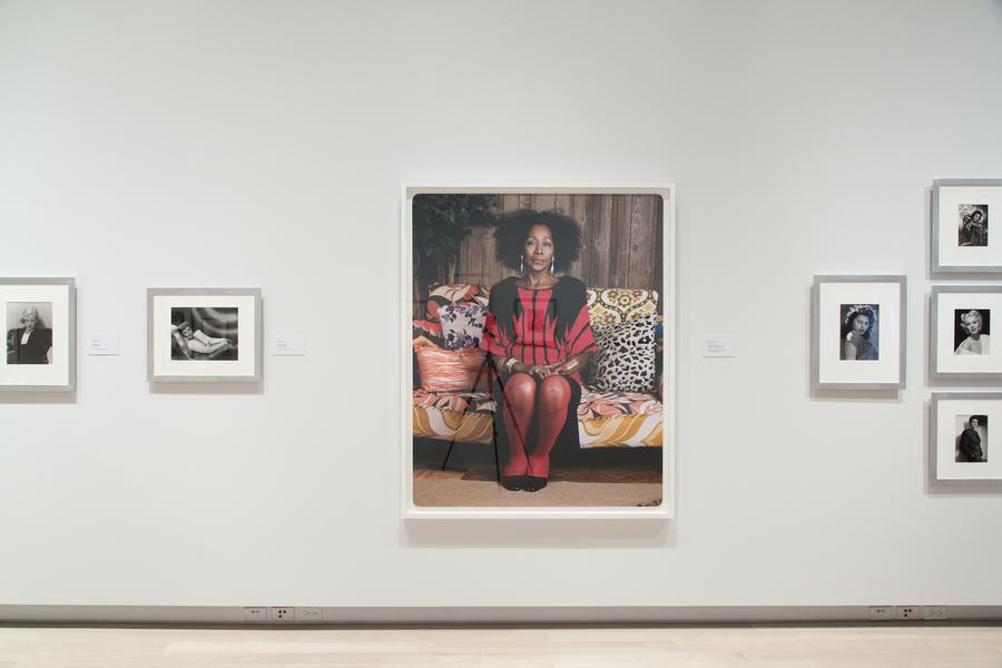 Large portrait of a woman on a brightly coloured couch, smaller black and white photographs framed on the wall beside it
