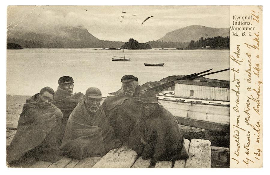 A group of men sit under blankets on a wooden dock, with the ocean and mountains in the background