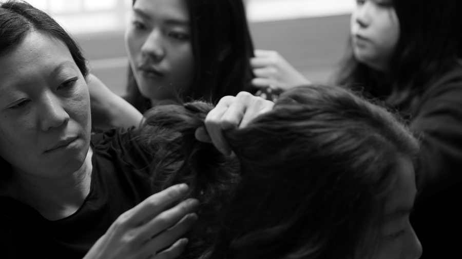 Four East Asian woman braid each others hair in a circle, each braiding the next closing the loop
