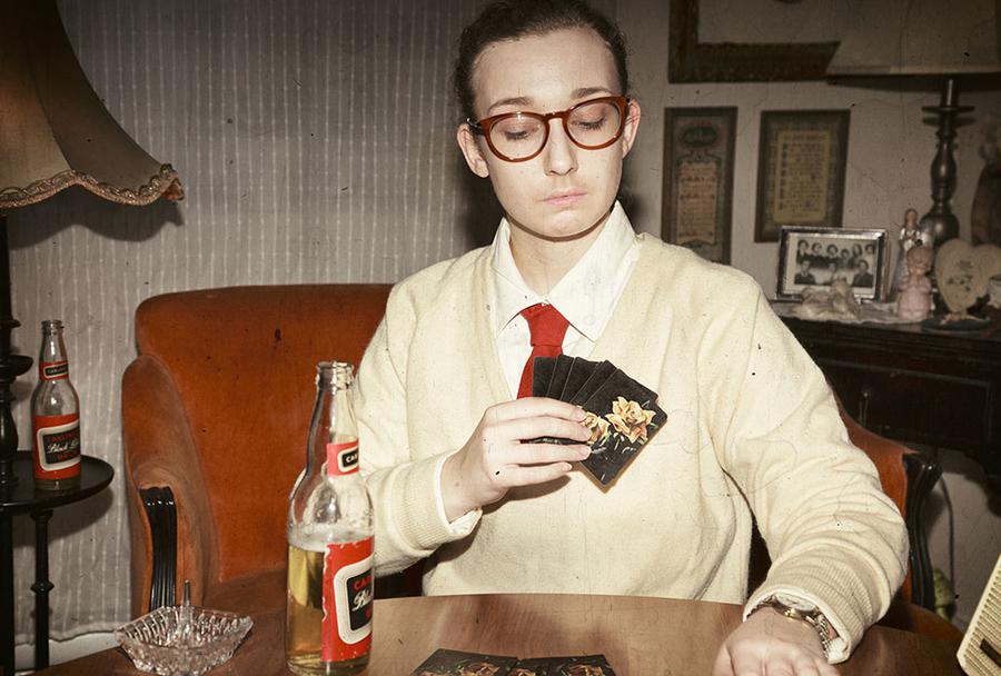 A person in a sweater and a tie looks down at a dining room table while holding a hand of cards, in a vintage-looking image.