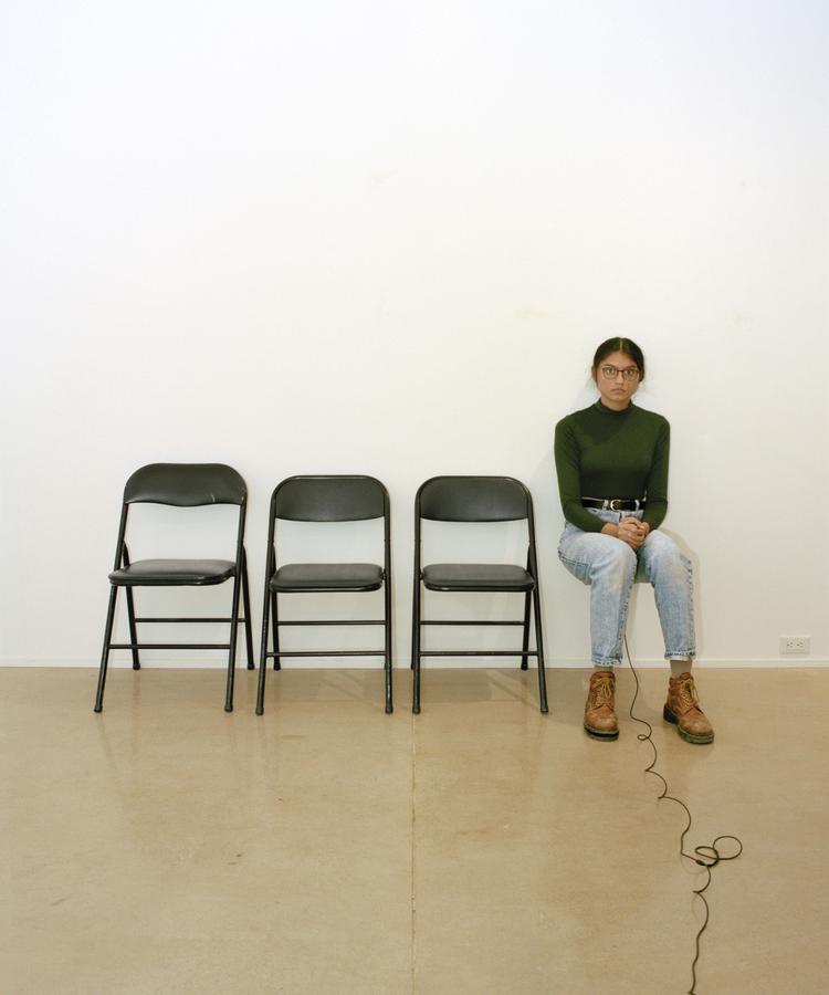 Woman in brown boots, blue jeans and green turtleneck sits with arms in her lap holding a microphone with the cord extending forward out of frame. She sits on one of four black folding chairs lined up in a row.