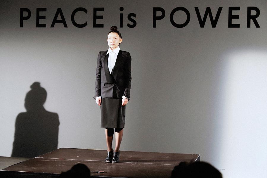 east asian woman in a black skirted suit and bunned hair stands on a platform in from of a wall with lettering that states "Peace is Power"