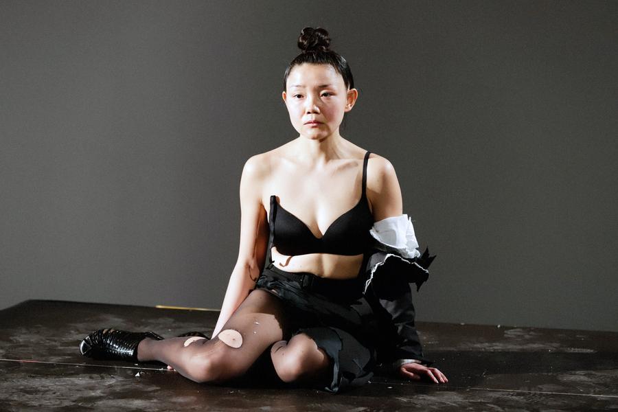 east asian women sits on platform stripped of shirt with bra strap slid off of one shoulder, tear in pantyhose and facial expression is desolate