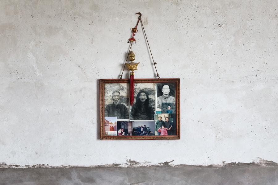 A frame holding family photographs hangs on a blank white wall, with a red and gold adornment.