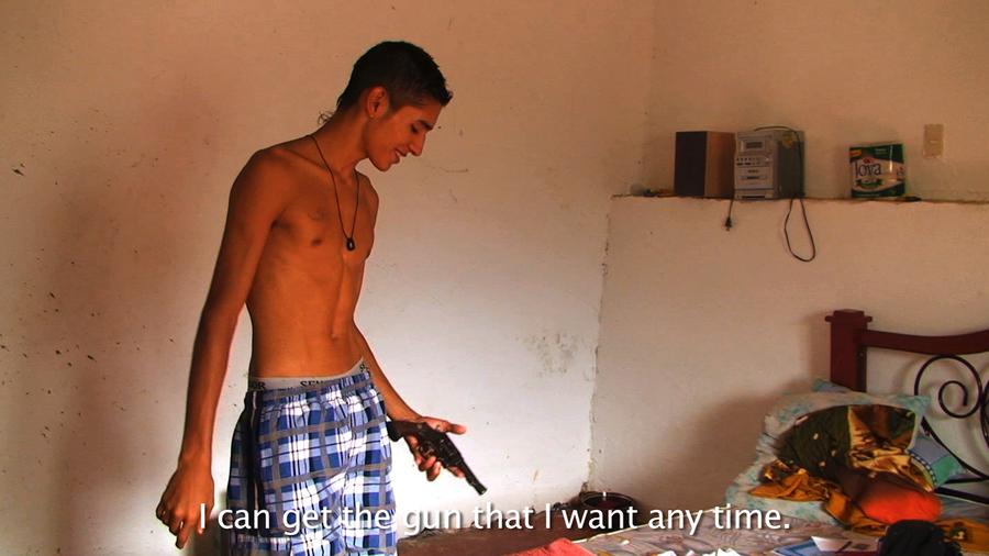 A shirtless young man wearing blue and white plaid shorts holds a gun. Text reads "I can get the gun that I want any time"