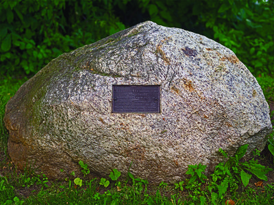 A large rock with a bronze historical plaque embedded into it is surrounded by grass.