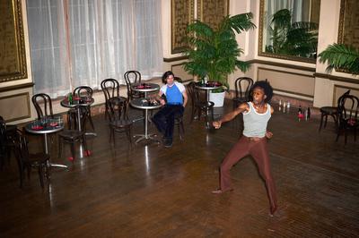 A man wearing red pants dancing in a large room, another man sits at a table and watches